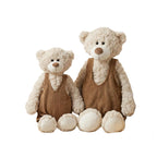 Two plush teddy bears in matching brown corduroy-style overalls posed together on a soft beige blanket with pillows and wooden toys in a warm nursery-style setting.