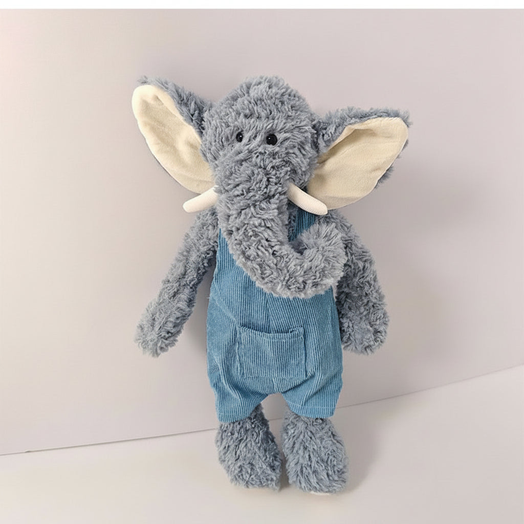 A plush elephant toy wearing blue corduroy-style overalls, photographed against a plain light background. The elephant has gray-blue fur, cream inner ears, white tusks, and a curly trunk.